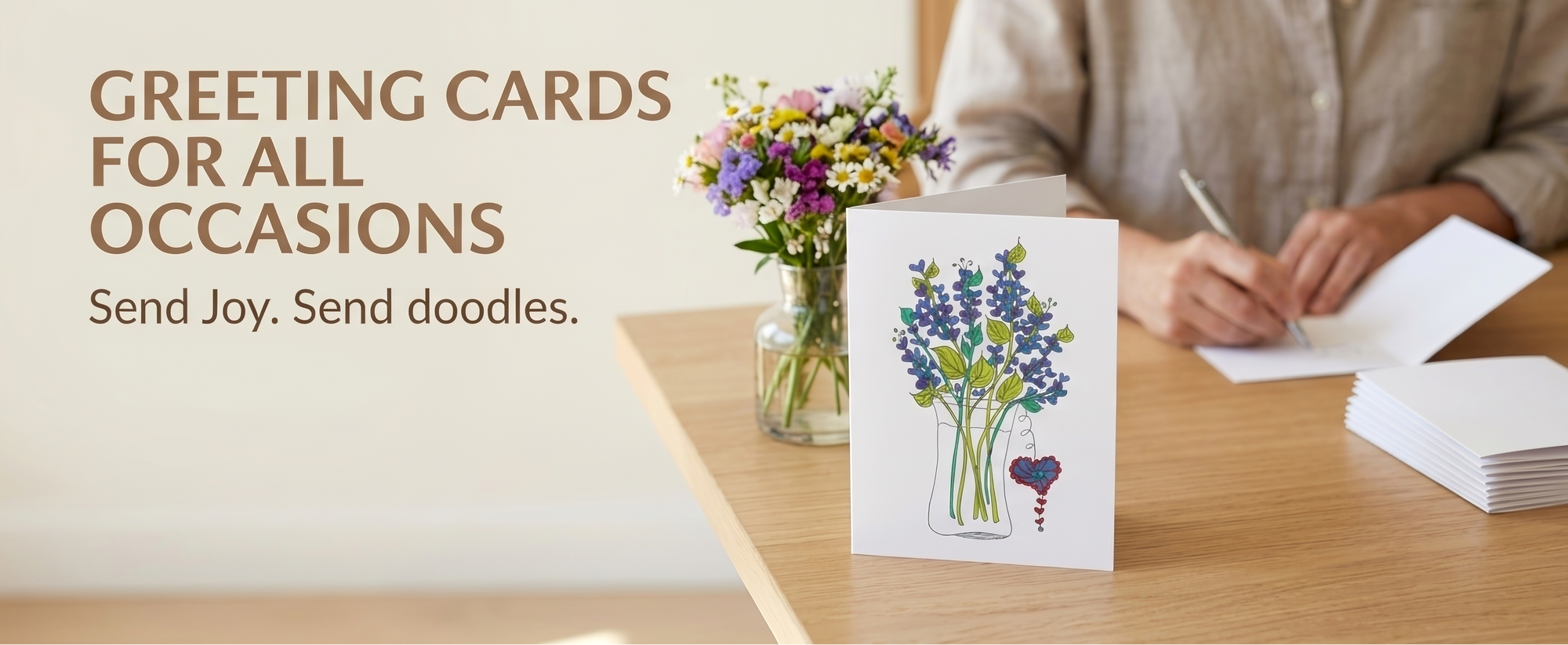 A greeting card with a blue and purple flower illustration stands on a wooden table next to a small vase of flowers. A person in a beige shirt is seated in the background writing in a notebook.
